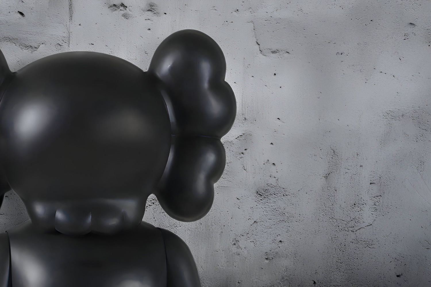 A large, dark gray sculpture with rounded shapes resembling an abstract figure stands against a textured, light gray concrete wall. The figure faces away with two ear-like protrusions.