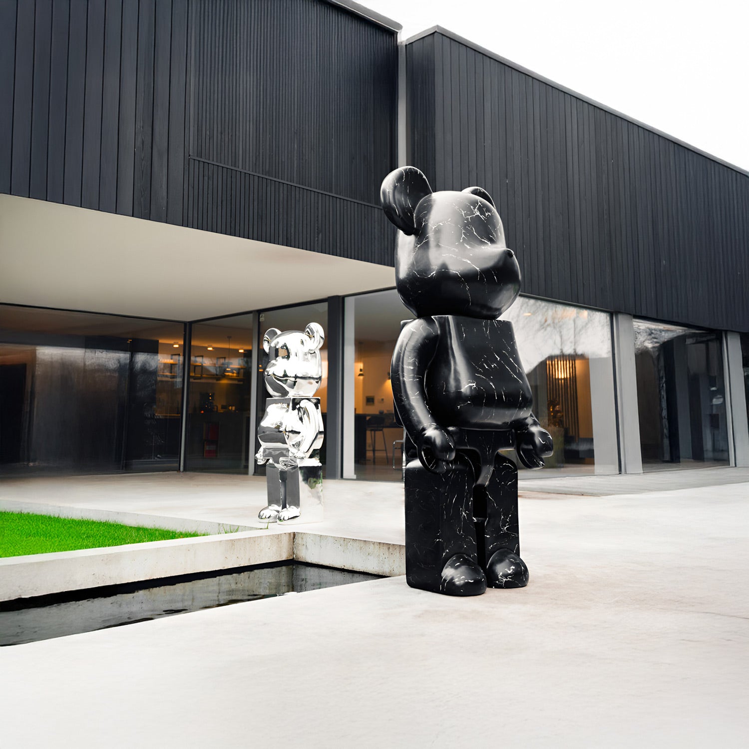 Two large bear sculptures, one black and one silver, stand outside a modern building with dark panels and large windows. The black sculpture is closer, set near a grassy rectangular patch and a narrow water feature.