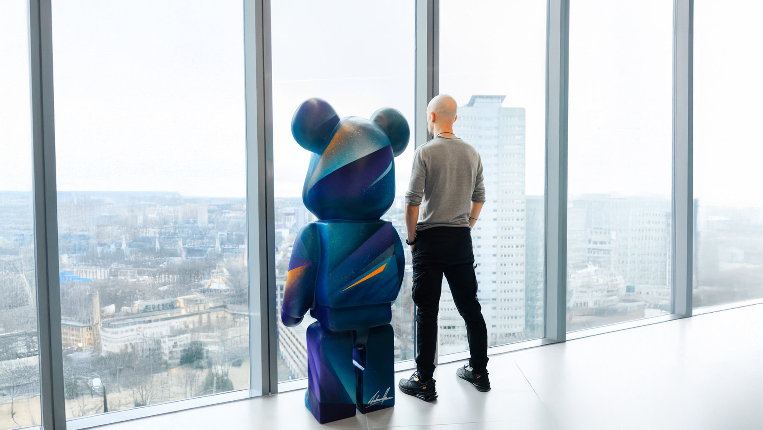 A man stands next to a large colorful bear sculpture, both gazing out of a high-rise window with a panoramic city view in the background.