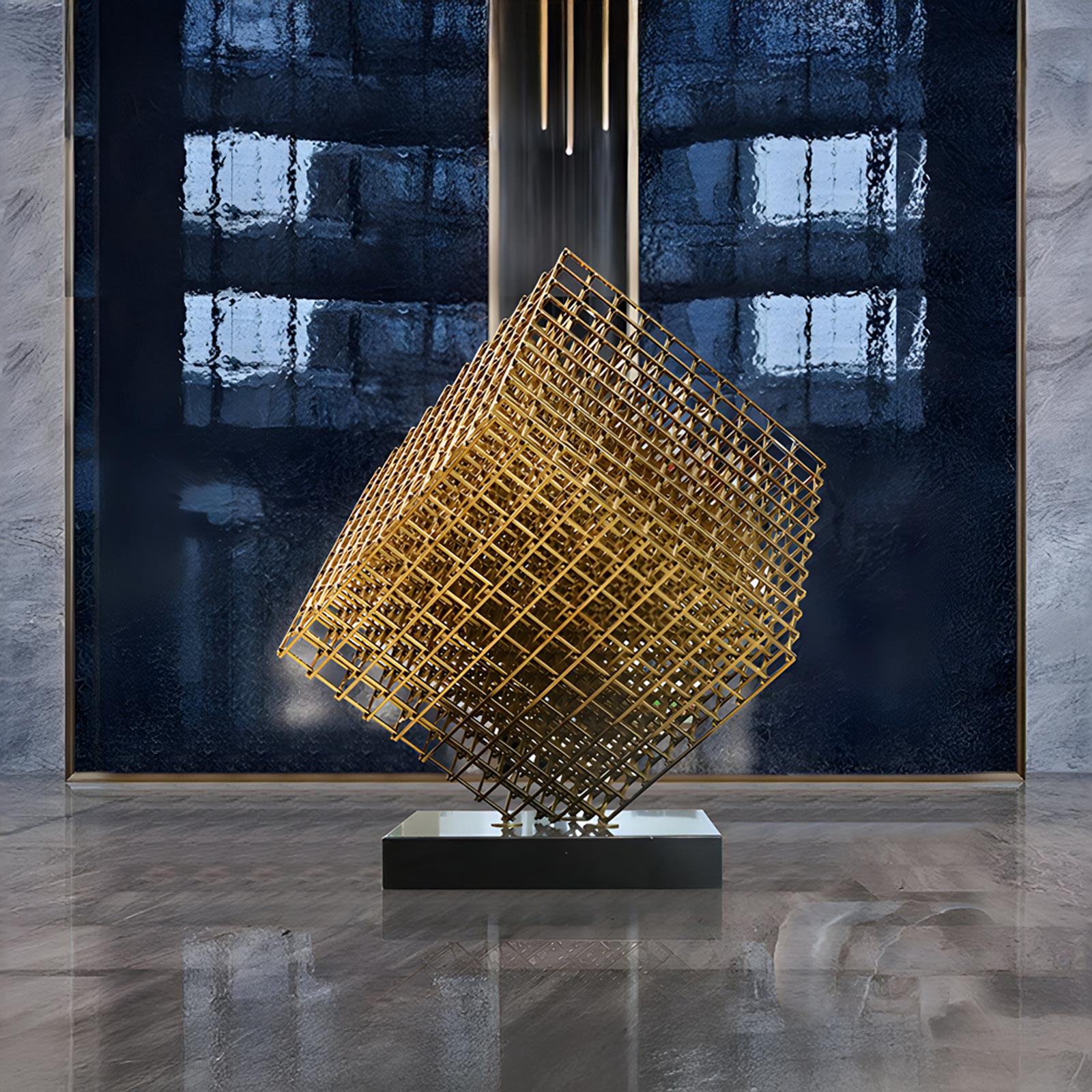 A modern stainless steel geometric sculpture featuring a gold-toned lattice cube balanced on one corner atop a sleek black base. The sculpture is set against a reflective dark blue glass wall in a contemporary interior with polished marble flooring, illuminated by vertical pendant lights above.