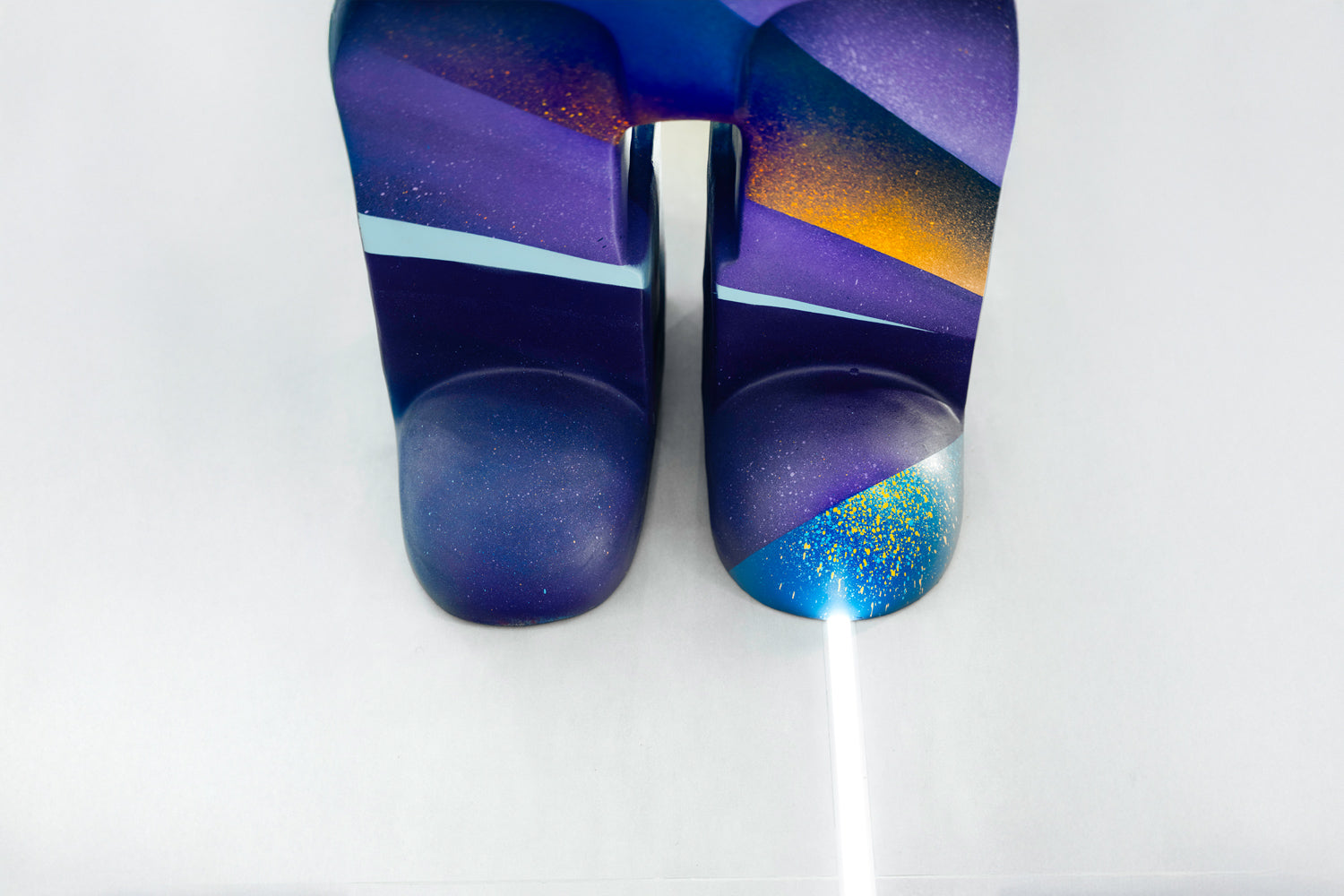 A colorful sculpture shaped like two abstract legs is painted with vibrant shades of purple, blue, and orange. The artwork stands on a glossy surface, with a thin beam of light pointing towards it from the bottom.