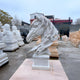 The 100cm Dynamic Horse Head Marble Outdoor Sculpture by Giant Sculptures gracefully sits on a pedestal amidst other yard sculptures. Its detailed flowing mane and expressive eyes form an elegant scene, complemented by surrounding buildings and leafless trees.
