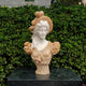 The Classical Lady Marble Bust - 65cm by Giant Sculptures, featuring wavy hair, a hat with a large bow, and a ruffled dress, is shown on a dark stone pedestal in front of dense green bushes.