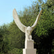 The Swooping Arc Large Owl Outdoor Sculpture by Giant Sculptures, standing 150cm tall in hand-carved marble, depicts a sleek owl in motion. It is displayed on a pedestal amidst lush greenery under the sky.