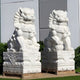 Two large Chinese Guardian Lion II sculptures by Giant Sculptures, intricately hand-carved in marble with detailed manes and standing 120cm high, adorn square pedestals outdoors. Positioned in front of a building amid lush greenery, these statues offer timeless elegance.