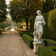 Flora Goddess of Spring 160cm white marble sculpture by Giant Sculptures, holding a flower basket on a pedestal beside a manicured hedge in a formal garden.