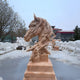 Under a cloudy sky, the Fluid Motion Horse Head Marble Outdoor Sculpture by Giant Sculptures, standing 100cm tall, exudes equine elegance with its flowing mane on a pedestal. It is surrounded by other stone sculptures amidst trees and buildings, creating a perfect outdoor backdrop.