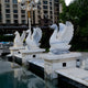 Giant Sculptures Wings-Lifted Swan II Marble Outdoor Sculptures, each 120cm tall, rest gracefully on square pedestals by a reflective pool. Behind them stands a modern apartment building with trees, and above is an elegant decorative lamppost.