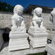 Giant Sculptures Chinese Guardian Lion III Marble Outdoor Sculptures (200cm) sit on ornate pedestals, featuring detailed manes and fierce expressions to enhance your outdoor décor against a white brick wall under a clear blue sky.