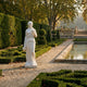 Standing Woman with Cherubs 160cm white marble sculpture by Giant Sculptures, holding a bowl, displayed on a gravel path beside manicured hedges and a reflecting pool.