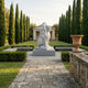 A stone pathway leads to the Giant Gracious Lion Marble Outdoor Sculpture - 120cm by Giant Sculptures, set on a pedestal amid manicured hedges, benches, and cypress trees, with a classical pavilion in the background.
