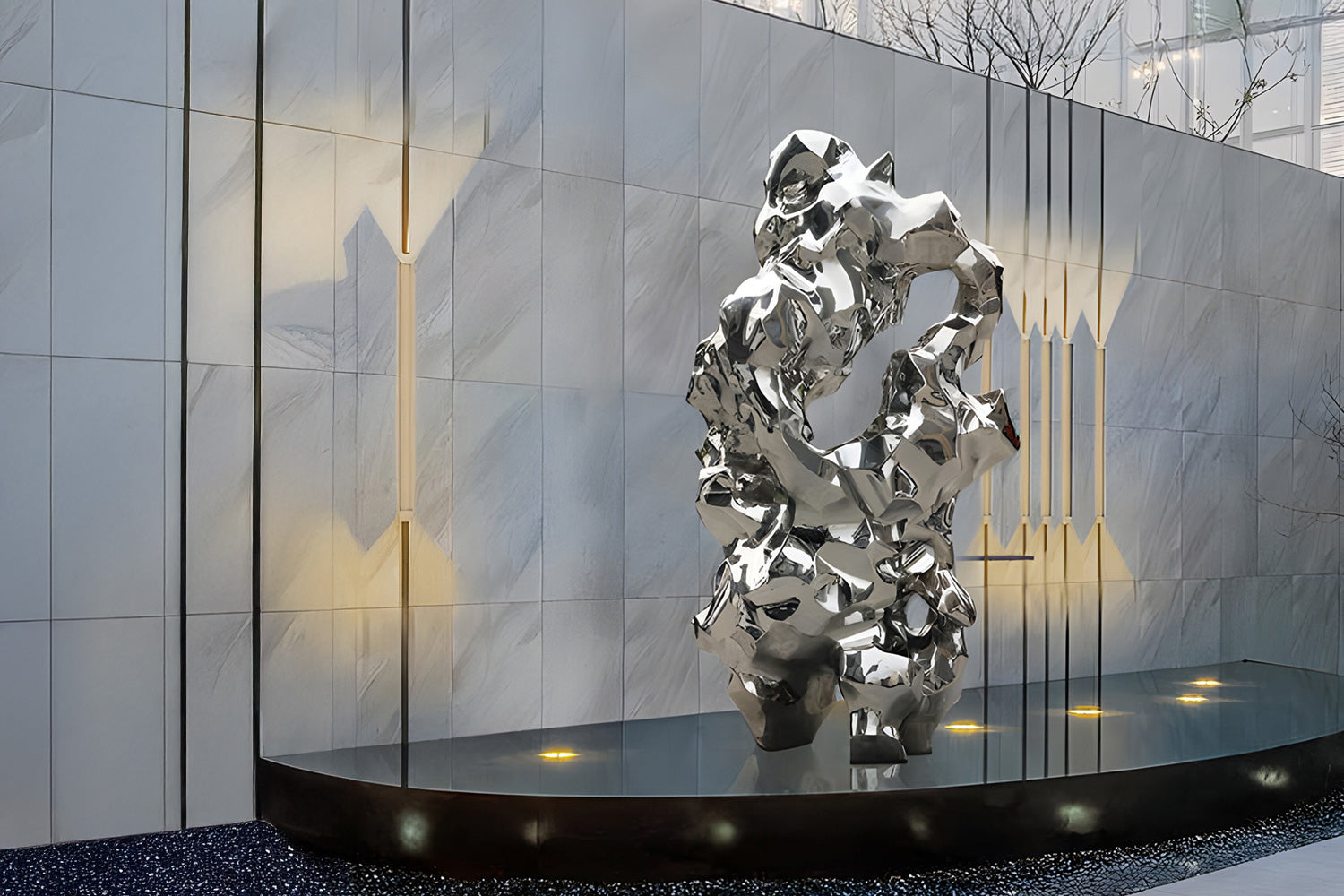 A modern abstract sculpture made of reflective silver metal stands on a circular platform. It is positioned against a textured wall with subtle vertical lighting accents. The setting appears to be an outdoor or semi-enclosed urban space.