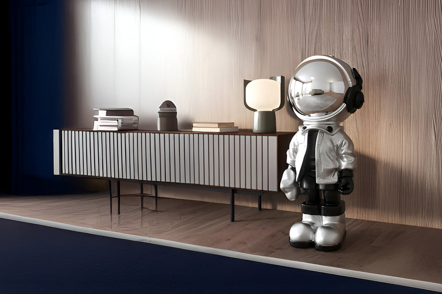 A modern living room scene featuring a large astronaut figurine in shiny attire beside a minimalist sideboard. The sideboard holds stacked books, a small lamp, and other decor, all set against a wooden-paneled wall.