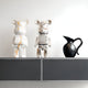 A pair of bear-shaped sculptures grace the shelf, one being the Mystic Chrome Bear Sculpture by Giant Sculptures, standing 50cm tall and adorned with geometric patterns, while the other displays a mirrored finish. They accompany a black ceramic pitcher featuring a curved handle against the backdrop of an unadorned white wall.