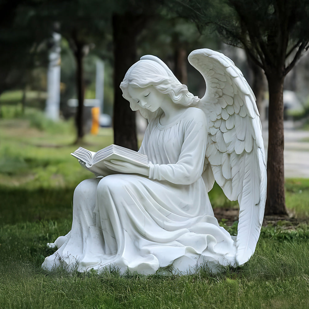 A white marble statue of an angel with wings, seated on grass, reading a book. The figure is surrounded by trees, creating a serene and peaceful atmosphere.