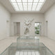 The Angelic Embrace Marble Outdoor Sculpture (260cm) by Giant Sculptures stands at the center of a minimalist, sunlit gallery with glass cases and large windows revealing lush greenery outside.