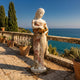 Caryatid Column Marble Sculpture 160cm by Giant Sculptures, a draped female figure holding roses, displayed on a seaside stone terrace with cypress trees and potted plants.