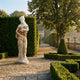 Caryatid Column Marble Sculpture 160cm by Giant Sculptures, a draped female figure holding roses, displayed on a gravel path in a hedged formal garden.