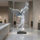 The Dove Release Peace Angel Marble Outdoor Sculpture - 280cm by Giant Sculptures, featuring a white marble angel with outstretched wings holding a dove aloft, is displayed on a pedestal among other exhibits in a spacious, modern gallery.