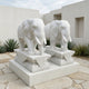 Two Lucky Chinese Elephant II Marble Outdoor Sculptures by Giant Sculptures, each 180cm tall, stand side by side on decorative pedestals amid geometric stone tiles and desert plants beside a modern white building.