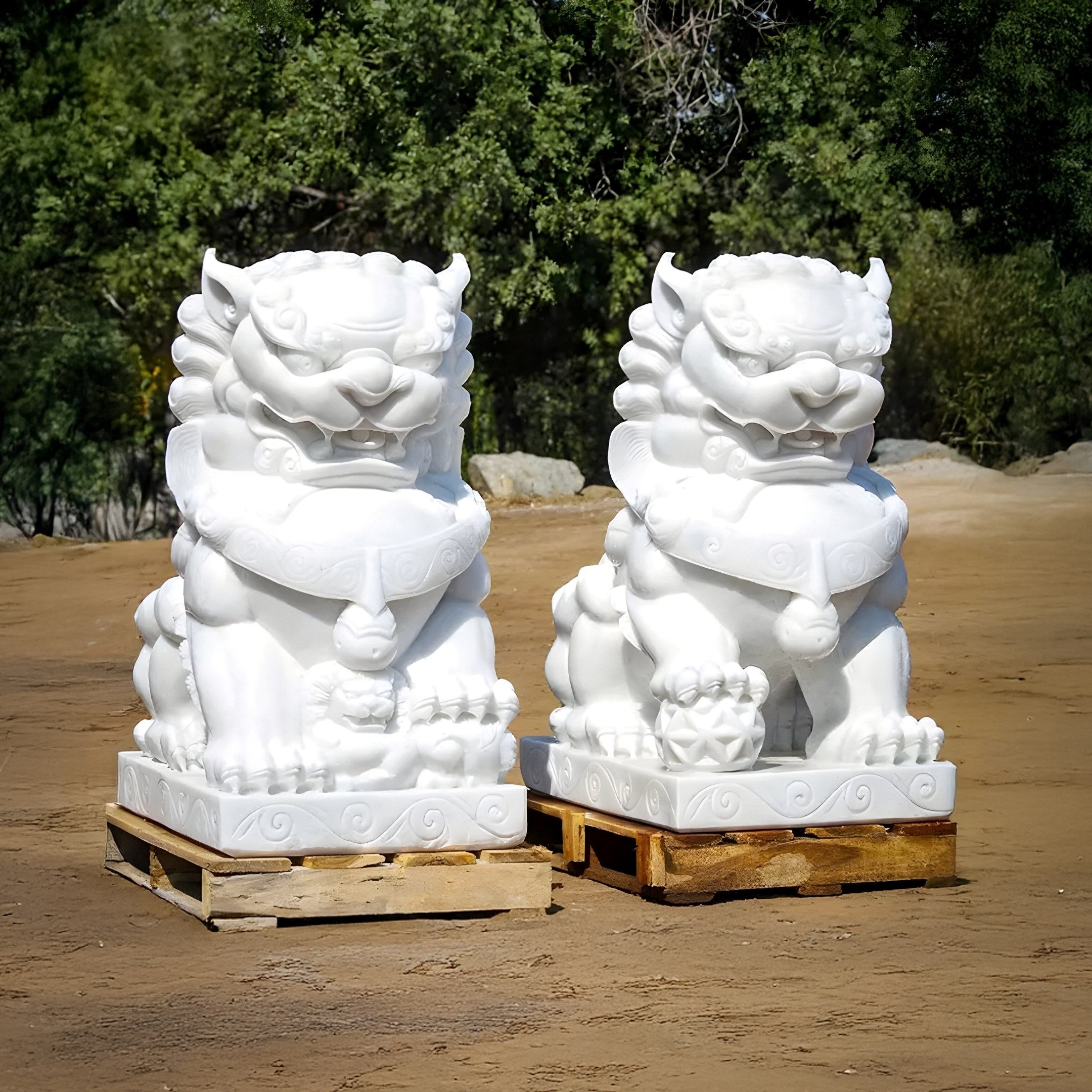 The captivating Giant Sculptures Chinese Guardian Lion VII marble outdoor statues (120cm) sit on wooden pallets amid lush green trees, showcasing intricate carved details.