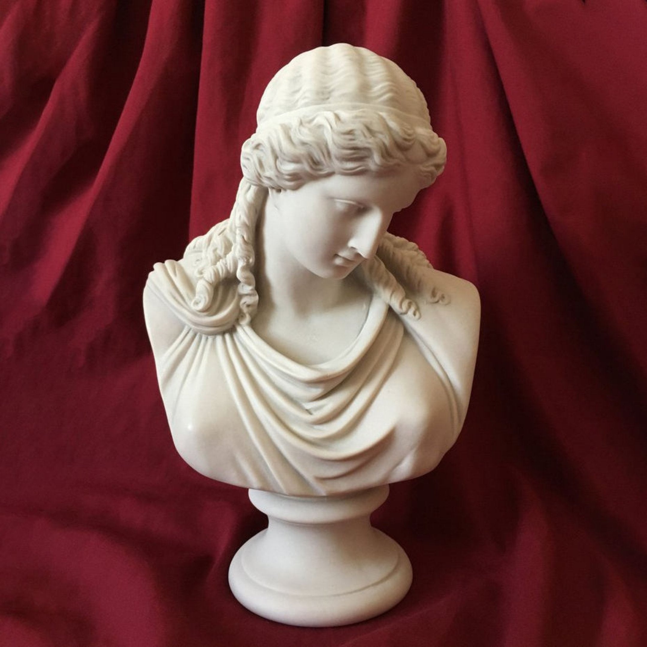 The Classical Beauty Marble Bust Sculpture - 65cm by Giant Sculptures features a woman with wavy hair and draped clothing, gazing downward, set against deep red, gathered fabric.