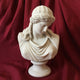 The Classical Beauty Marble Bust Sculpture - 65cm by Giant Sculptures features a woman with wavy hair and draped clothing, gazing downward, set against deep red, gathered fabric.