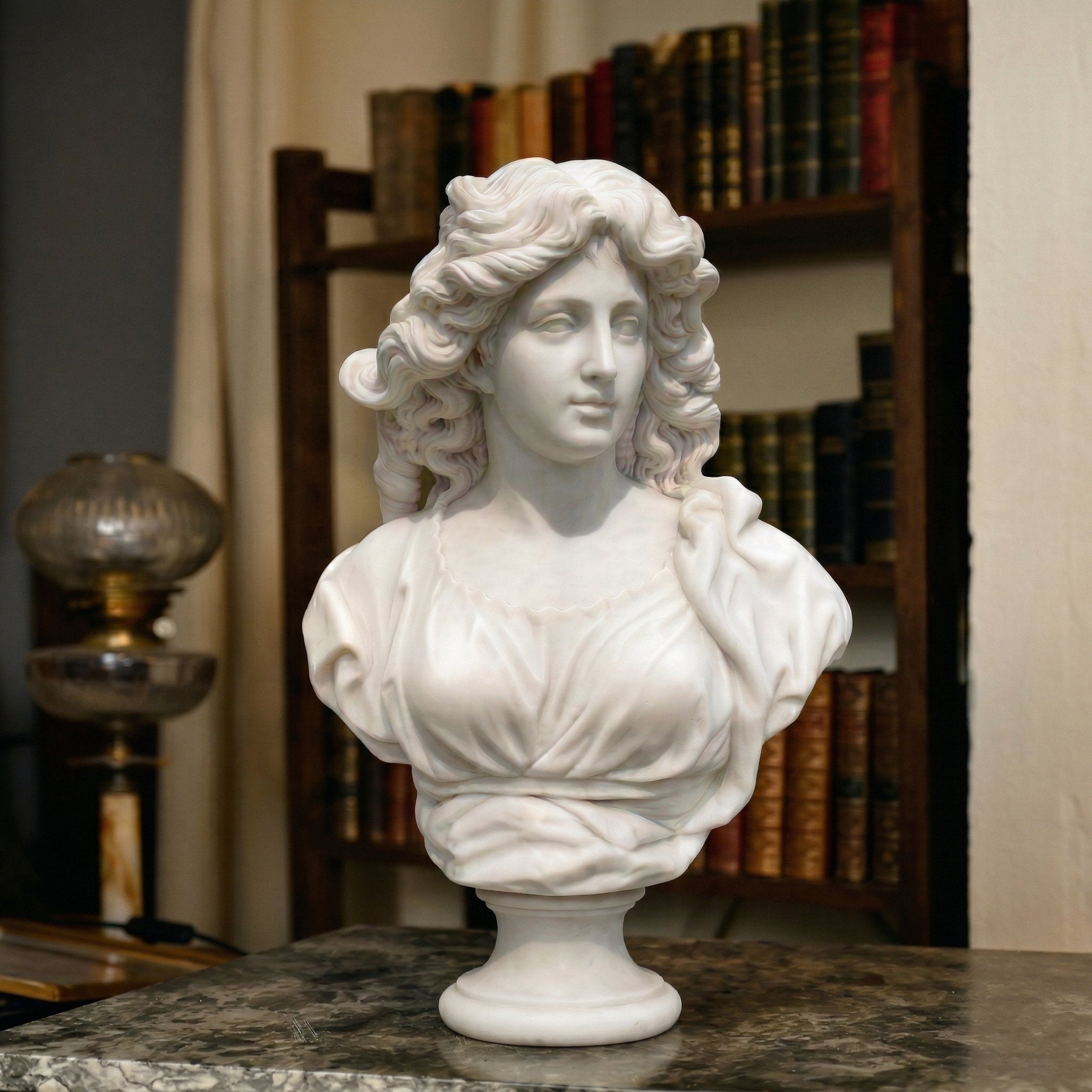 The Classical Female Marble Bust Sculpture - 65cm by Giant Sculptures features wavy hair and draped clothing, displayed on a white marble surface with bookshelves and an antique lamp in the background.