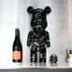 A Giant Sculptures Black Stone Bear Sculpture - 50cm, featuring a sleek water-transferred black stone finish, is displayed on a shelf next to a dark wine bottle adorned with an orange label. Nearby, a small black rounded object lies beside a stack of books against the backdrop of a silver-gray wall, crafting an elegant modern interior decor scene.