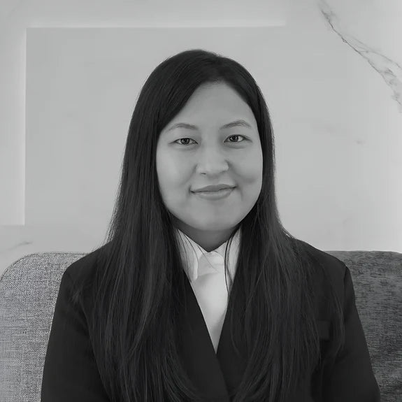 A woman with long dark hair wearing a suit and white shirt sits against a light-colored background. She is facing forward and smiling slightly.