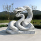 The Ferocious Snake White Marble Outdoor Sculpture (100cm) by Giant Sculptures, intricately carved and double-headed with open mouths, stands on a smooth paved surface against rolling hills and leafy trees beneath a clear blue sky.