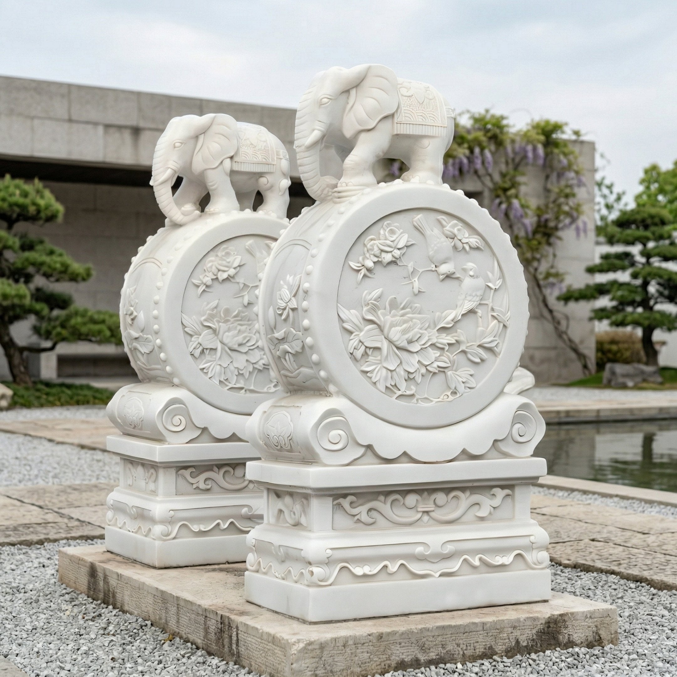 The Elephant On Drum Marble Outdoor Sculpture - 120cm by Giant Sculptures features hand-carved marble elephants with floral details on decorative pedestals, enhancing outdoor elegance by a pond and manicured trees.