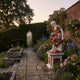 Giant Sculptures Seated Woman with Wreath polychrome marble sculpture on red and white pedestal, set in a misty walled garden with roses and lavender.