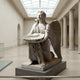 The Shell Offering Angel Marble Outdoor Sculpture - 150cm by Giant Sculptures features a kneeling angel with a shell-shaped bowl, beautifully displayed indoors beneath tall columns and a glass ceiling.