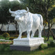 The Majestic Bull White Marble Outdoor Sculpture by Giant Sculptures stands elegantly on a decorated pedestal amidst a garden, featuring intricate details of the bulls muscular build, curved horns, and focused expression with lush bushes and trees providing a serene backdrop.