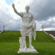 The Augustus Marble Sculpture - 180cm by Giant Sculptures depicts a man in Roman armor with one arm raised, standing on grass beside a small figure. Steps, torches, and a cloudy sky can be seen in the background.