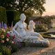 Reclining Goddess with Cherub and Flowers Marble Sculpture by Giant Sculptures, displayed on a stone terrace beside roses, lavender, and a lily pond at dusk.