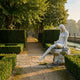 Seated Woman Marble Sculpture 130cm by Giant Sculptures resting on a stone block beside a hedged garden path, with trees and a still pond nearby.
