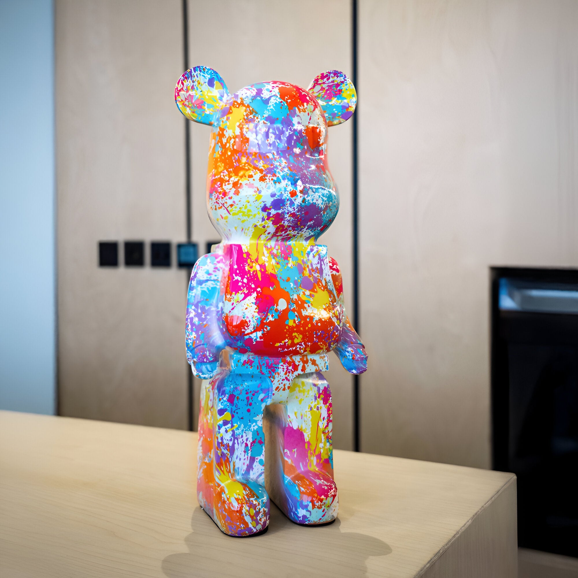 The Hype Bear Sculpture - 50cm by Giant Sculptures is a striking figure of modern design standing on a light wooden surface. Its exterior is adorned with dynamic, abstract splashes of blue, yellow, pink, orange, and purple paint that evoke the essence of urban street art. This vibrant piece contrasts beautifully against the backdrop of a minimalist interior with neutral tones.