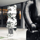 A towering Mystic Chrome Bear Sculpture - 135cm by Giant Sculptures, with a reflective silver form and chrome electroplated finish, stands on a concrete surface outside a modern building. In front, the blurred outline of another Mystic Chrome Bear Sculpture subtly captures modern design aesthetics.