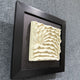 The Harmonia 3D Ceramic Wave Wall Sculpture by Giant Sculptures features layered cream ceramic waves in a textured, dimensional design set against a black frame, shown mounted on a gray carpeted surface.