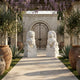 A stone pathway lined with potted plants leads to a wrought iron gate, flanked by Giant Sculptures’ Chinese Guardian Lion IV Marble Outdoor Sculpture (180cm). Purple flowers hang from a pergola as sunlight casts soft shadows on the tiled ground.