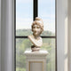 The Female Marble Bust With Cap Sculpture - 50cm by Giant Sculptures is displayed on a pedestal before a tall window, showcasing lush green countryside and trees outside.