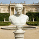 The Classical Male Marble Bust Sculpture - 75cm by Giant Sculptures is displayed on a pedestal in a formal garden with trimmed bushes, a circular fountain, and a grand classical building in the background.