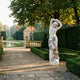 Standing Woman with Roses Marble Sculpture by Giant Sculptures, 160cm white figure with terracotta roses and sage leaves, displayed on a garden path beside a reflecting pool.