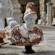 The Bearded Male Marble Bust Sculpture - 90cm by Giant Sculptures features an ancient warrior in a plumed helmet and armor, crafted from white and brown natural marble and displayed outdoors on a black pedestal among other statues.