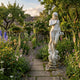 Giant Sculptures' 160cm Standing Maiden marble sculpture in flowing robes on a weathered pedestal, set along a stone pathway among foxgloves, roses, and lavender.