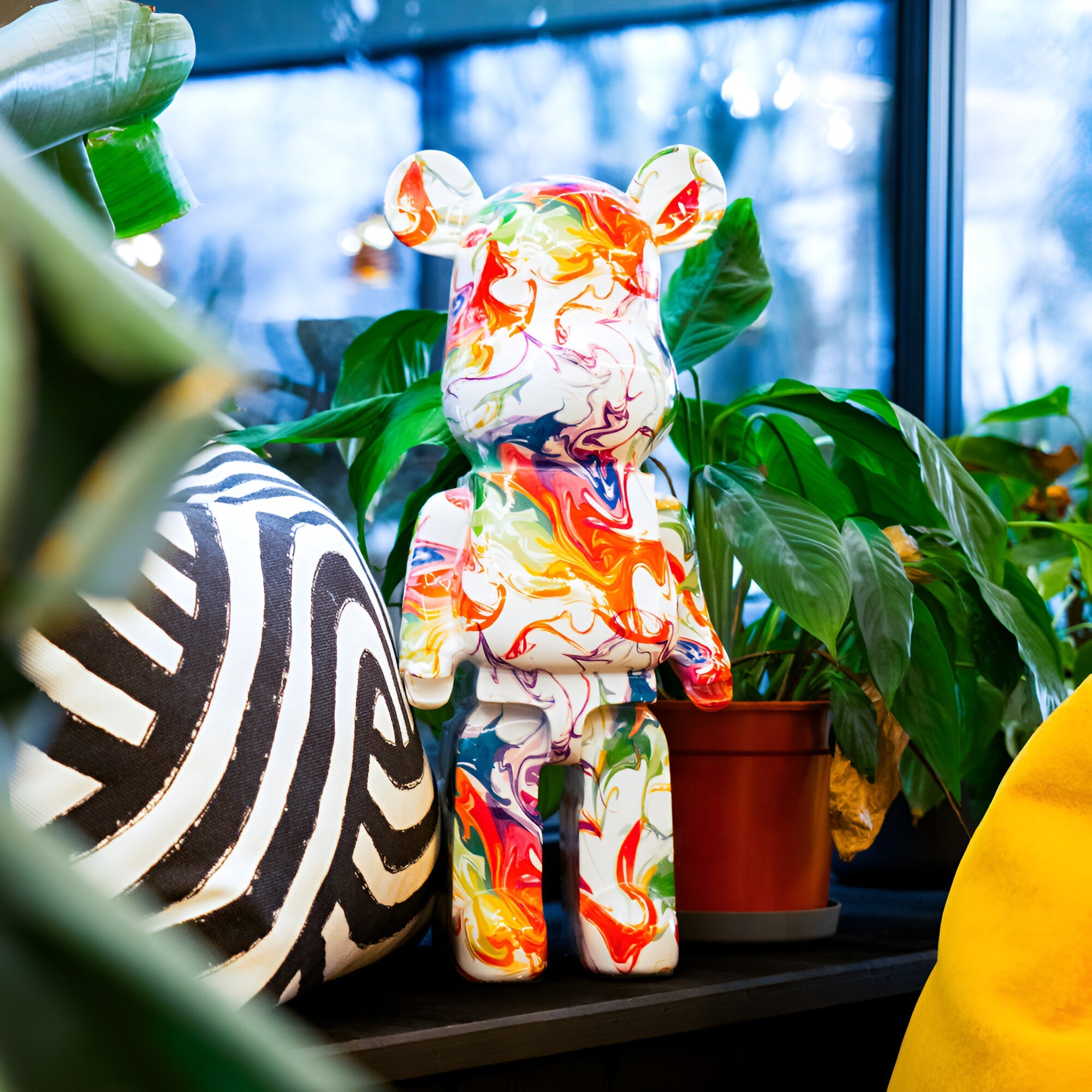 A vibrant Vogue Bear Sculpture from Giant Sculptures, featuring an abstract pattern and standing at 50cm tall, is showcased amidst lush green plants. To the left is a large black-and-white patterned item, while a brown potted plant adorns the right side. The contemporary design scene unfolds against a window with softly blurred outdoor views.