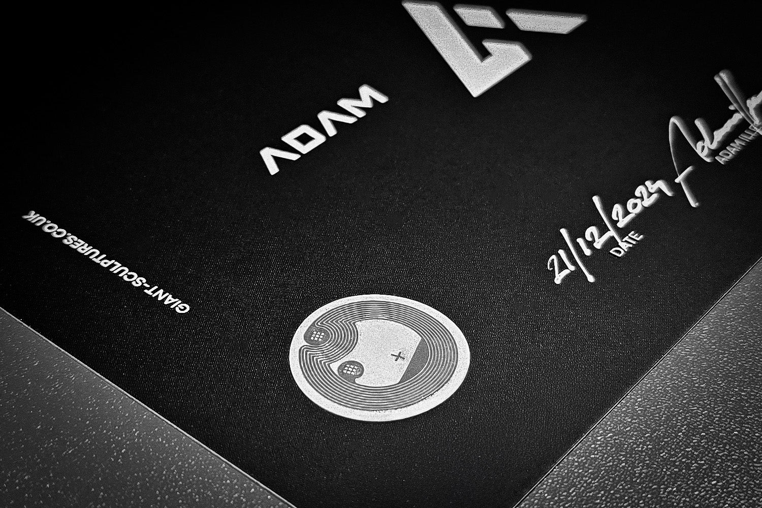 Close-up of a black certificate featuring a circular yin-yang symbol with a plus sign and a wireless charging icon. The certificate includes white text, a date 21/12/2024, a signature, and branding elements. The surface has a subtle texture.