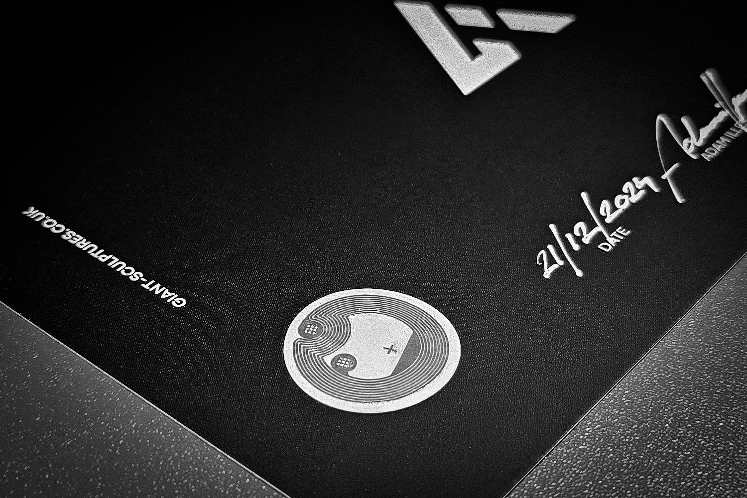 A black card featuring a silver yin-yang symbol with plus and minus signs. The date 21/12/2023 and a signature are visible. The corner features part of a geometric logo and the text GIANT-SCOURGE.CO.UK.